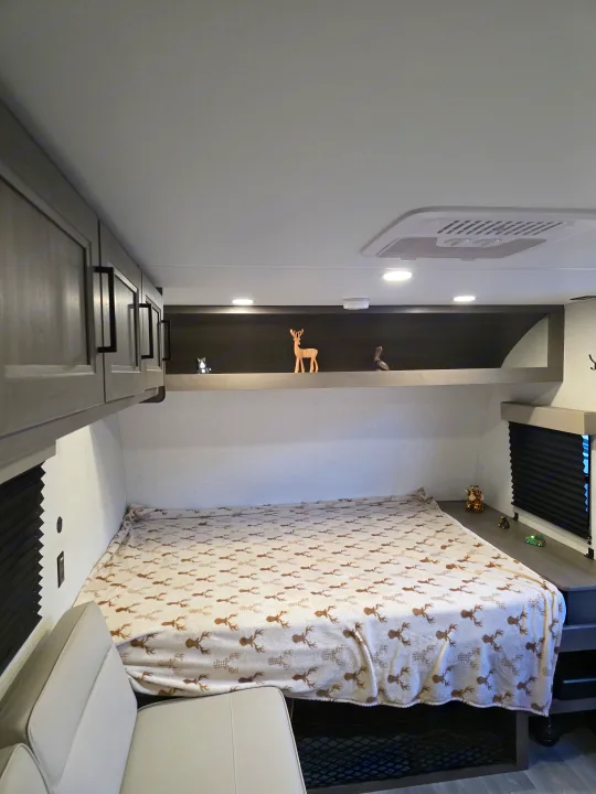 Inside a comfortable RV rental bedroom, featuring a bed with deer print blanket, storage, and decorative shelf with figurines.. Grand Design Transcend One 2025