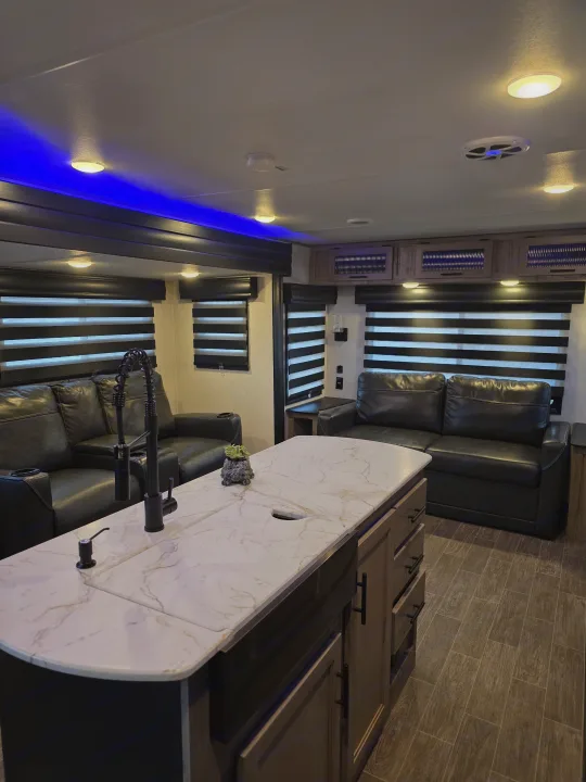 Modern RV rental interior featuring a kitchen island with a black faucet, black leather seating, horizontal blinds, and blue accent lighting.. Forest River Cherokee Alpha Wolf 2022