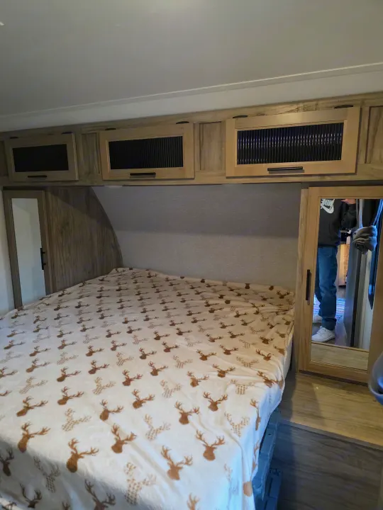 Cozy RV rental bedroom features a bed with a deer blanket, overhead cabinets, and a mirrored closet reflecting a person.. Forest River Cherokee Alpha Wolf 2022