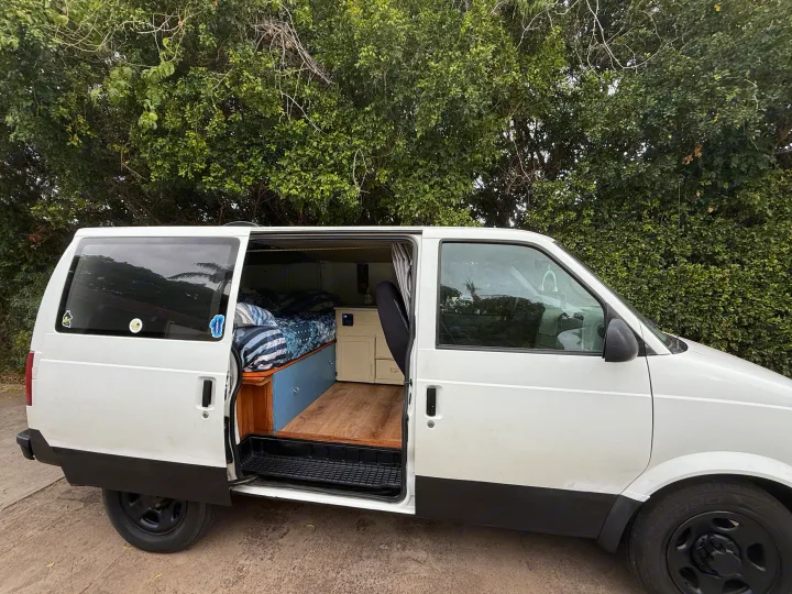 A white converted van for RV rental, side door open showing a bed, wooden floor, and storage inside. Lush trees in background.. Chevrolet Other 2005
