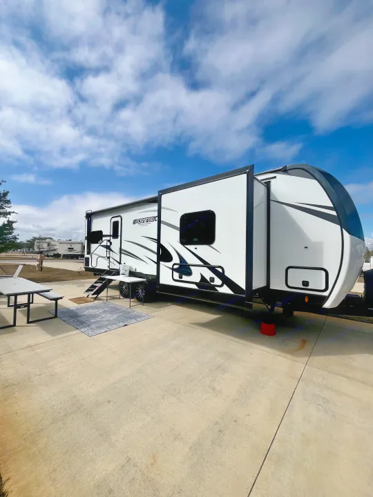 White and black RV rental set up on a concrete pad with outdoor furniture at a campground under a blue sky.. Sporttrek Touring Edition 2024