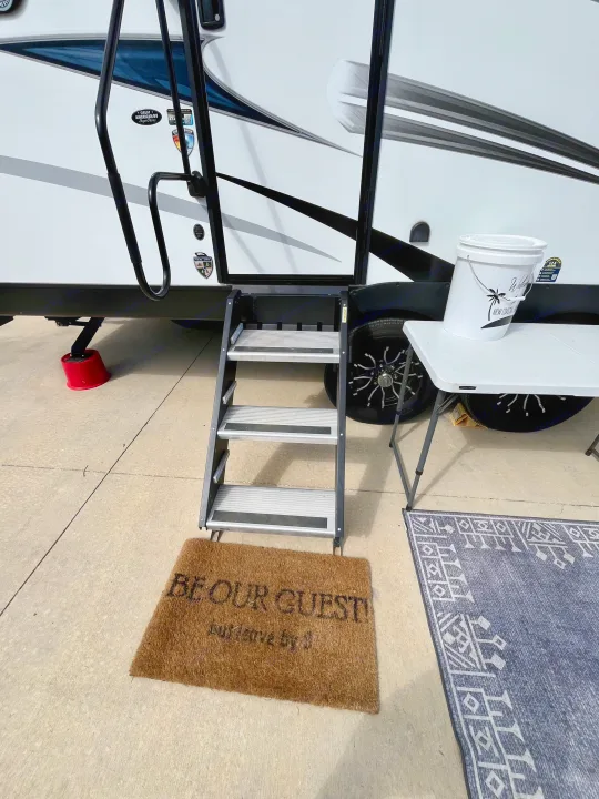 RV rental steps welcome guests with a "BE OUR GUEST!" doormat, outdoor table, and bucket, ready for a cozy stay.. Sporttrek Touring Edition 2024