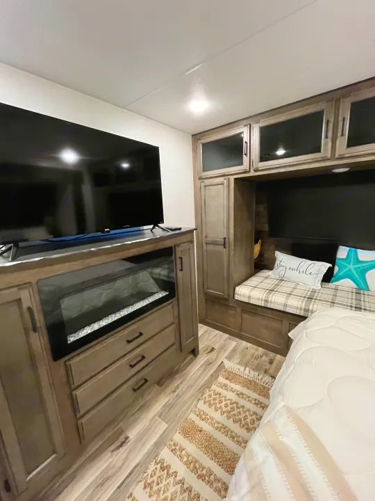 Inside a modern RV rental: a cozy bedroom with a large TV, electric fireplace, wood-look cabinets, padded bench, bed with white comforter.. Sporttrek Touring Edition 2024