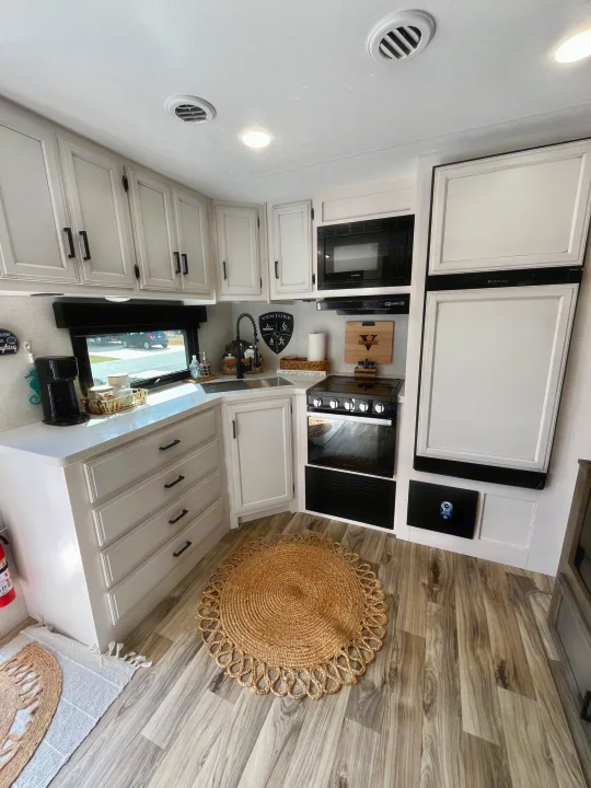 Modern RV rental kitchen featuring light cabinets, black appliances, white counters, and a jute rug for a comfortable stay.. Sporttrek Touring Edition 2024