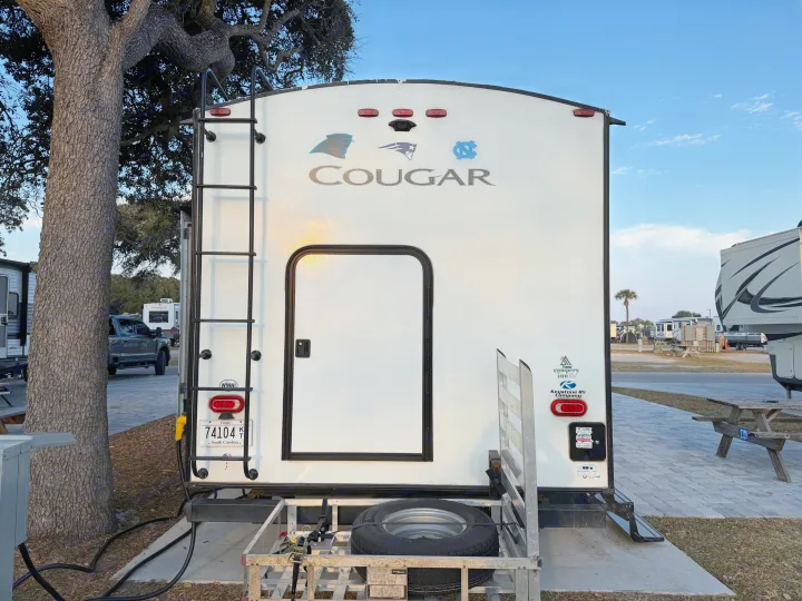 Back of a white Cougar rv rental, decorated with team logos, parked at a South Carolina campsite with a cargo carrier.. Keystone Cougar Half-Ton 2020