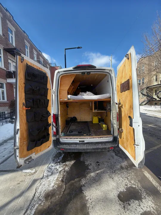Inside an RV rental van: open rear doors reveal a custom wooden interior, bed, storage, and a skateboard, parked on a snowy street.. Ford Transit 2021