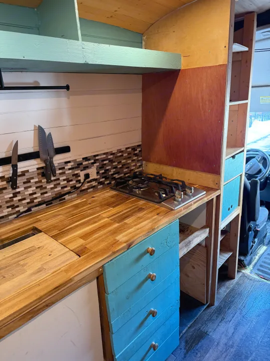 Cozy RV rental kitchen with wood counters, built-in gas stove, sink, magnetic knife strip, and charming blue cabinets.. Ford Transit 2021