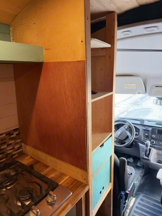 RV rental: DIY camper van interior features a kitchen area with stove, wood counter, and custom shelving, leading to the driver's cab.. Ford Transit 2021