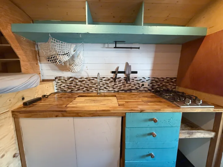 Inside this cozy RV rental, discover a compact kitchen with wood counters, sink, stovetop, blue drawers, and a sleeping area.. Ford Transit 2021