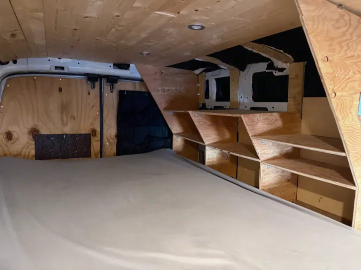 Inside an rv rental, this campervan features a wooden ceiling, comfy bed, and custom plywood storage shelves.. Ford Transit 2021