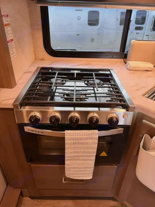 CONTOURE gas stove & oven in an RV rental kitchen, with a window view and a towel on the handle.. Thor Four Winds 2025