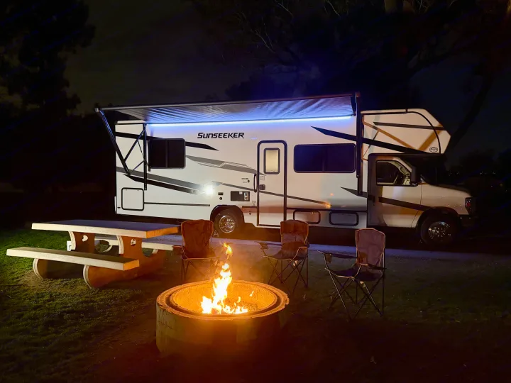 Cozy nighttime RV rental setup with a glowing fire pit, picnic table, chairs, and a "Sunseeker" RV under an illuminated awning.. Forest River Sunseeker LE 2025
