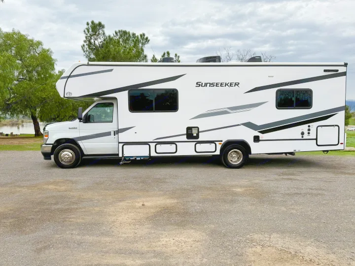 White Class C Sunseeker RV rental parked on a gravel lot, with a scenic backdrop of water and trees under a cloudy sky.. Forest River Sunseeker LE 2025