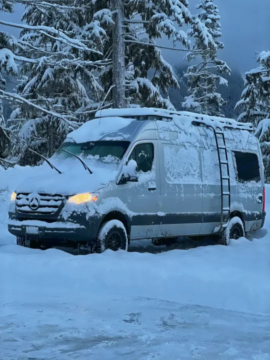 This snowy Mercedes Sprinter rv rental van, with headlights on, is nestled in a winter wonderland forest. Ready for cold-weather travel.. Mercedes-Benz Sprinter 2020