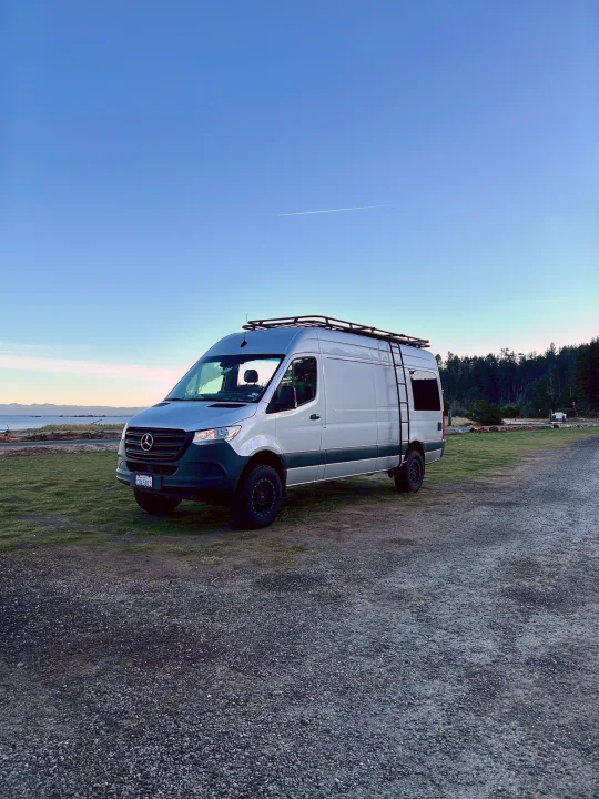Experience van life with this silver Mercedes Sprinter rv rental parked beside a serene coastal landscape and forest.. Mercedes-Benz Sprinter 2020