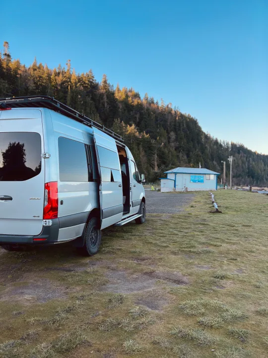 Silver Sprinter camper van (rv rental) with roof rack and open door, parked by a forest and 'Ocean Park Camp & Kayak' building.. Mercedes-Benz Sprinter 2020