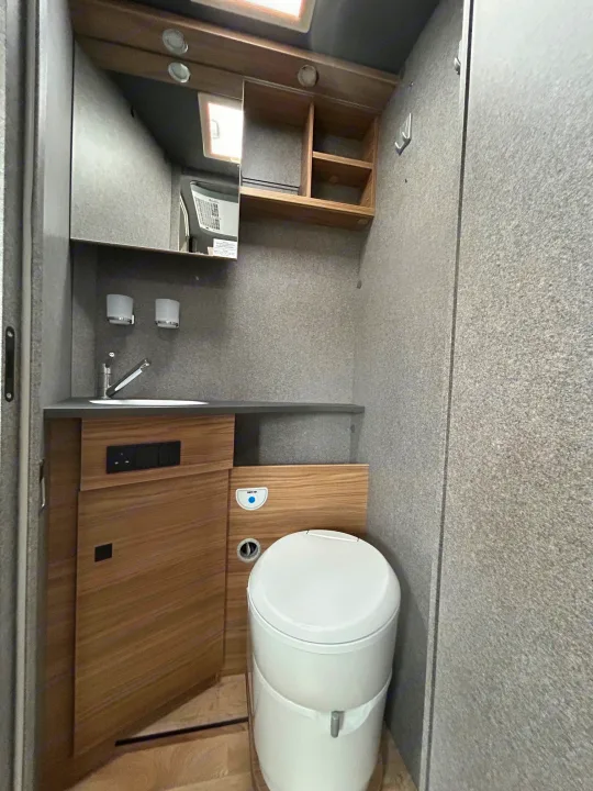 Compact RV rental bathroom featuring a toilet, sink, mirror, wooden storage, and power outlets for travelers.. GlamperRV DBL 2025