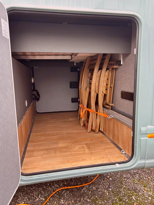 Spacious RV rental storage area with wooden flooring, holding folded chairs secured by an orange strap, ready for travel.. GlamperRV DBL 2025