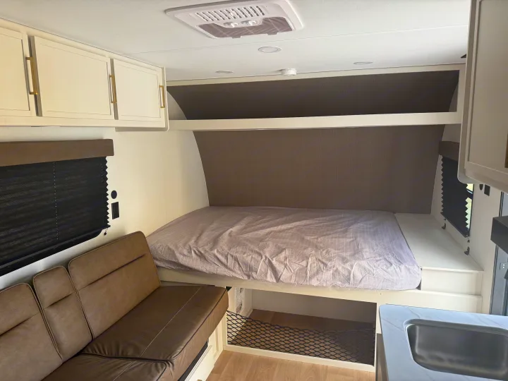 RV rental interior: cozy bed, brown sofa, overhead cabinets with gold handles, window blinds, and partial kitchen sink. Modern and compact.. Transcend One 151BH 2026