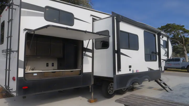 This white RV rental features an open outdoor kitchen with a sink, mini-fridge, and cabinets, ready for your next adventure.. Grand Design Reflection 2022