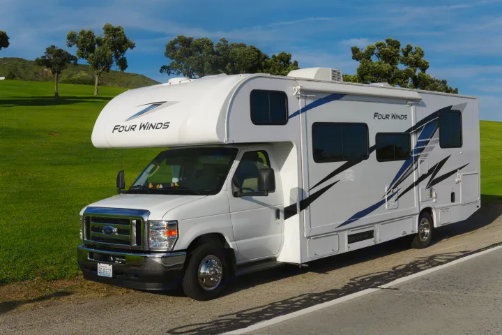A pristine white Four Winds Class C rv rental on a Ford chassis, parked by grassy hills under a blue sky.. Thor Four Winds 2025