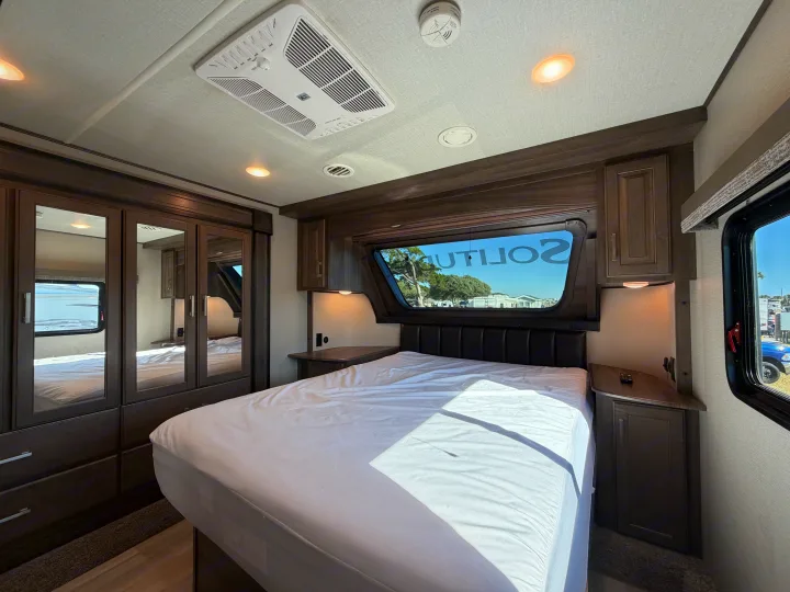 Modern RV rental bedroom featuring a comfy bed, dark wood storage, mirrored wardrobe, AC, and a scenic window view.. Grand Design Solitude 2021