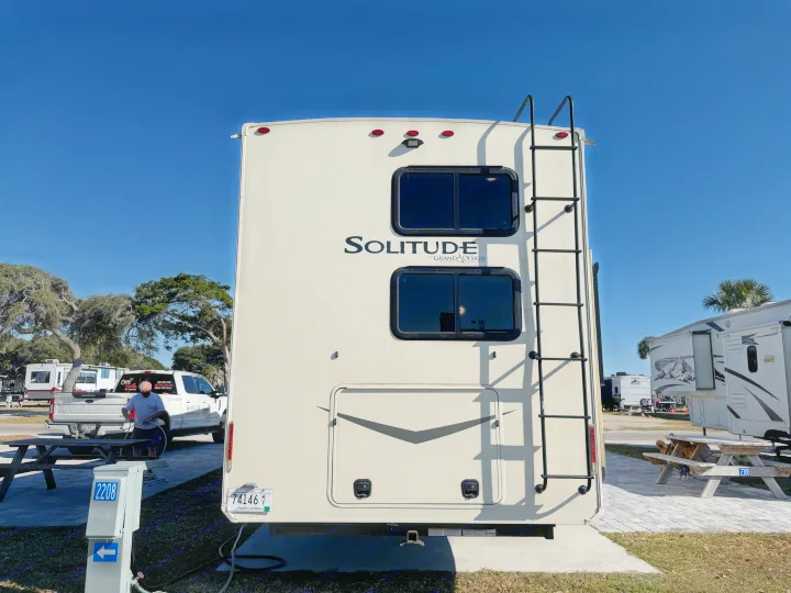 Solitude RV rental at a sunny campsite, showing its rear with a ladder, and a Cozy Turtle RV Rentals truck in the background.. Grand Design Solitude 2021