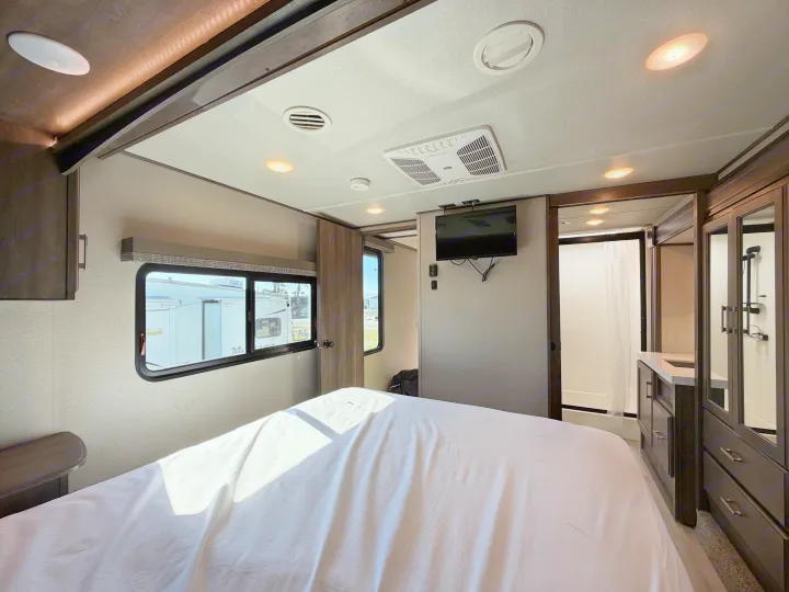 Interior of an RV rental bedroom with a large bed, sunlit windows, wall-mounted TV, and a glimpse of the ensuite bathroom.. Grand Design Solitude 2021