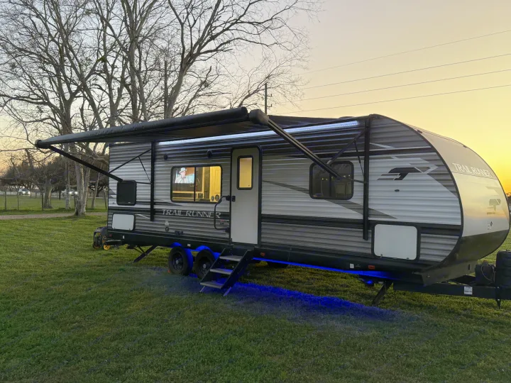 This Trail Runner RV rental glows with awning and undercarriage lights at sunset, TV visible inside, ready for a cozy camping evening.. Heartland RVs Trail Runner 2022