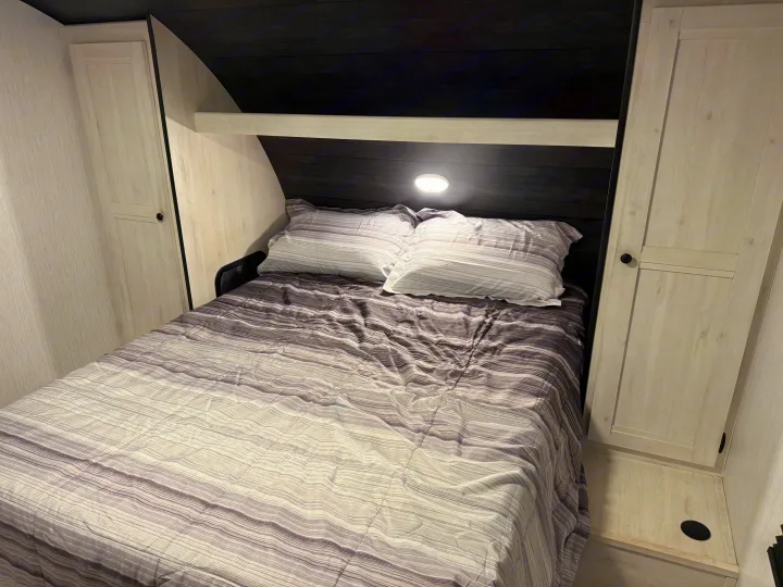 Cozy RV rental bedroom with a striped comforter, pillows, built-in cabinets, and a bedside light. Ideal for travel accommodation.. Heartland RVs Trail Runner 2022