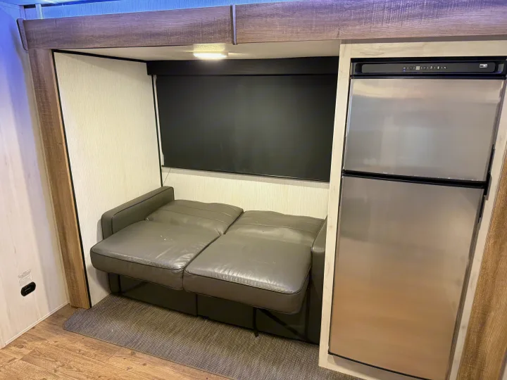 An RV rental interior showing a compact bunk space with a gray convertible sofa, dark window blind, and stainless steel fridge.. Heartland RVs Trail Runner 2022