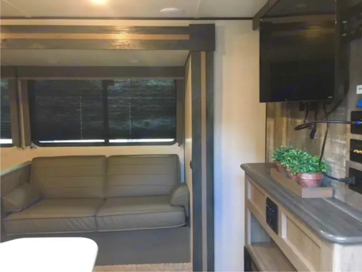 Inside an RV rental, a cozy living area with a grey sofa, window blinds, wall-mounted TV, and wood-toned entertainment console.. Keystone RV Hideout 2021