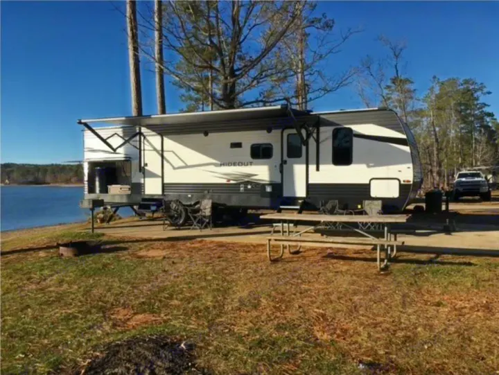 This RV rental is parked lakeside with an awning extended, a picnic table, and a fire pit, perfect for a scenic outdoor getaway.. Keystone RV Hideout 2021
