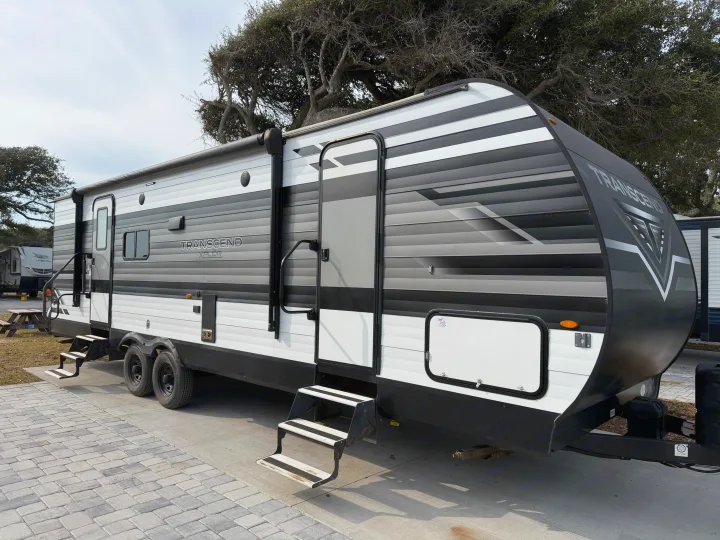 A gray and white Grand Design Transcend XPLOR travel trailer, an RV rental, is parked at a sunny campsite.. Grand Design Transcend 265BH 2022