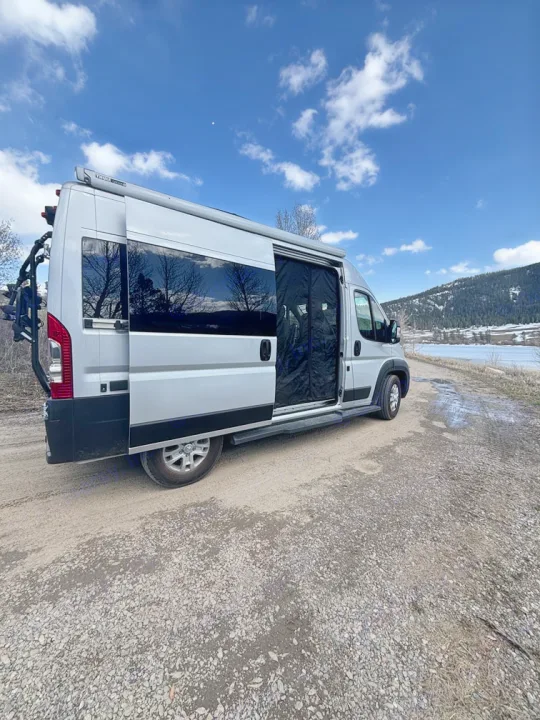Silver RV rental parked on a gravel road by a scenic lake and mountains, ideal for an adventure road trip.. Thor Scope 2024