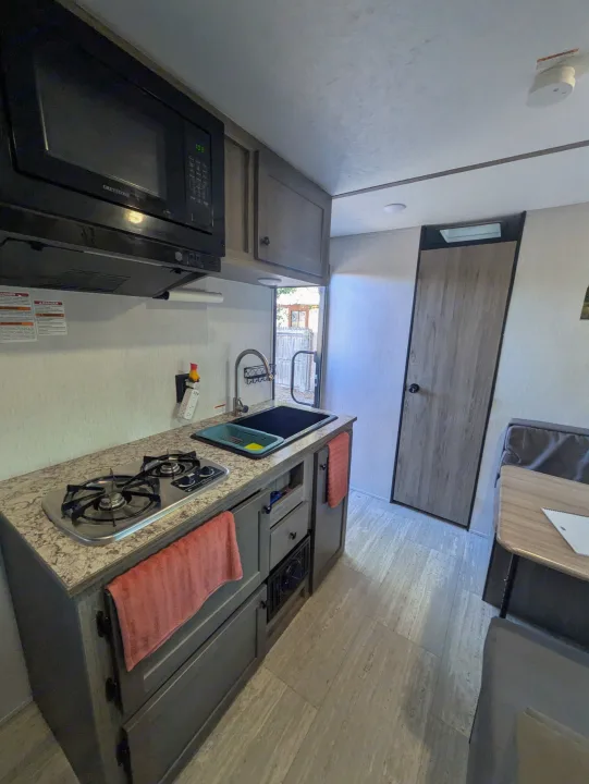 This RV rental's compact kitchen features a microwave, 2-burner stove, sink, and dining booth to the right.. Sunset Park & Rv Inc. 19rb 2025