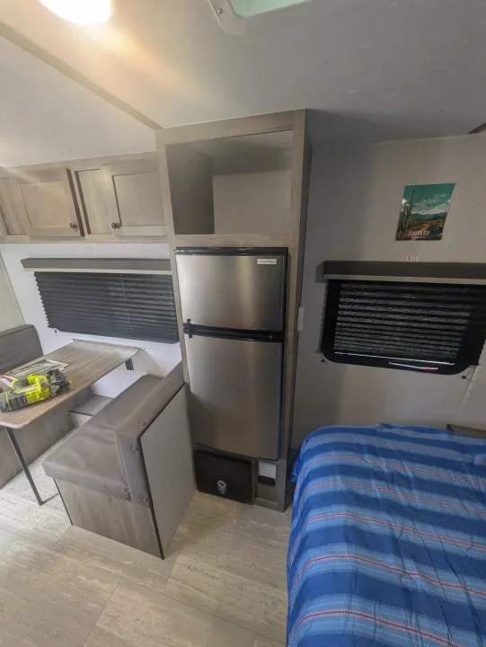 This RV rental features a compact kitchen/dinette area, fridge, storage, and a comfy bed with a striped comforter.. Sunset Park & Rv Inc. 19rb 2025