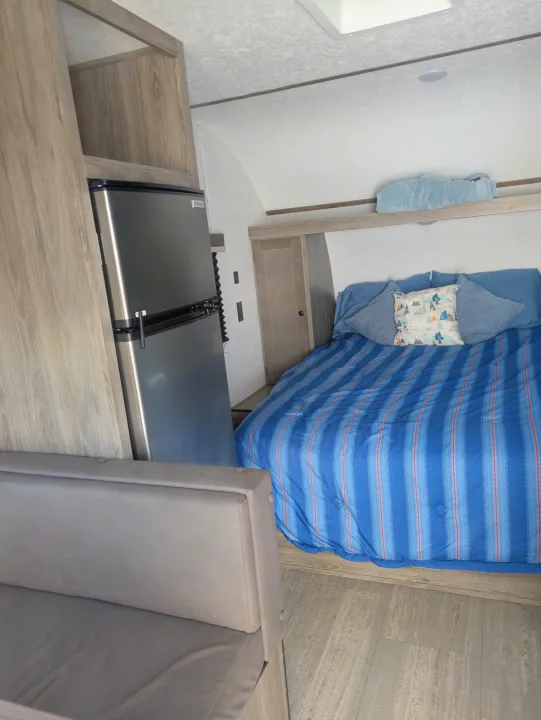 This cozy rv rental interior features a bed with blue bedding, a compact refrigerator, and a grey bench seat.. Sunset Park & Rv Inc. 19rb 2025