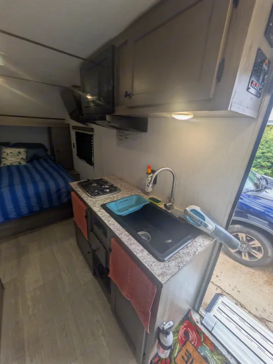 Cozy RV rental interior: compact kitchen (sink, stovetop, microwave) & comfy bed. Ideal for road trips & adventure.. Sunset Park & Rv Inc. 19rb 2025