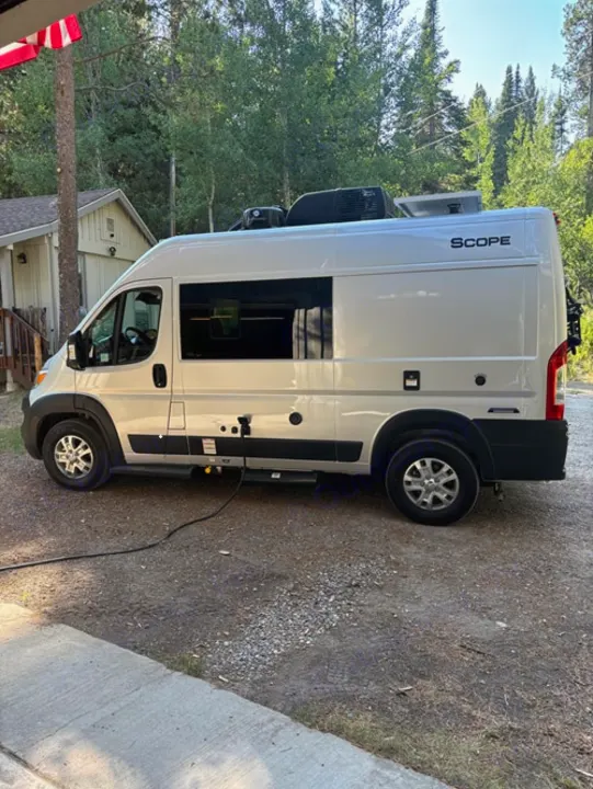 This white RV rental, a Scope camper van, is parked with a power hookup in a wooded area.. Thor Scope 2024