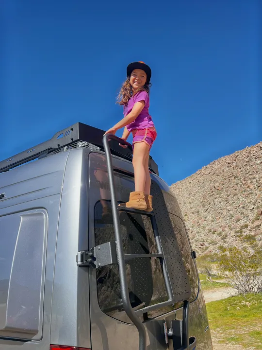 Joyful girl on a Winnebago Revel rv rental ladder, smiling under a blue desert sky. Perfect for family adventures!. Winnebago Revel 2025