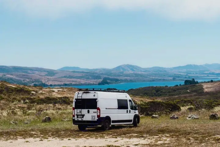 Your next rv rental adventure awaits! White camper van parked by a serene water body, surrounded by rolling hills under a blue sky.. Ram ProMaster 2022
