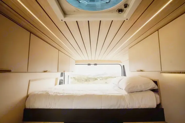 Experience van life! This modern RV rental interior features a cozy bed, wood ceiling with lighting, storage, and a scenic view.. Ram ProMaster 2022