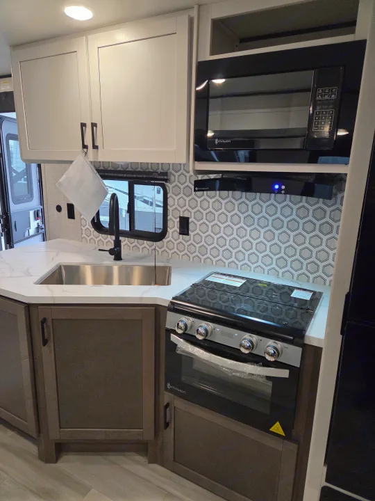 Modern RV rental kitchen showing sleek sink, black faucet, white countertops, microwave, and oven with hexagon backsplash.. Open Range Conventional 26BHS Travel Trailer 2025