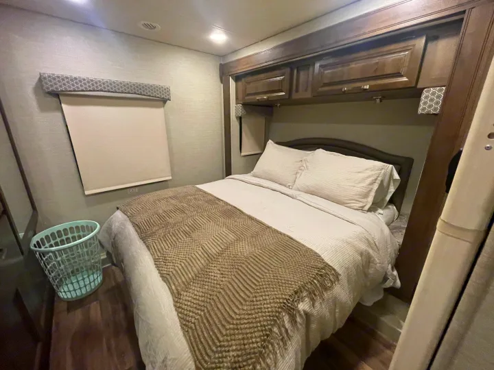Inside an RV rental: a neatly made bed with white linens & a textured throw, overhead storage, window shade, & laundry basket.. Jayco Greyhawk 2019