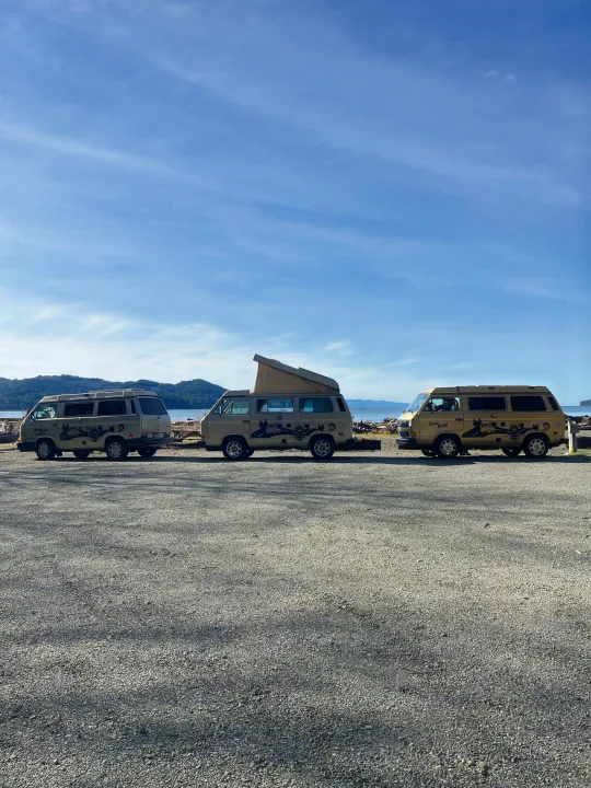 Three beige Westfalia RV rental camper vans, one with a pop-top, parked on a gravel lot by scenic mountains and water. Road trip ready!. Volkswagen Westfalia 1986