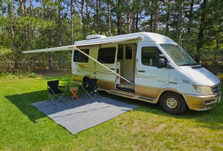 RV rental: White camper van with awning extended over outdoor seating and mat on green grass. Pine trees in background for camping.. Mercedes-Benz Sprinter 2005