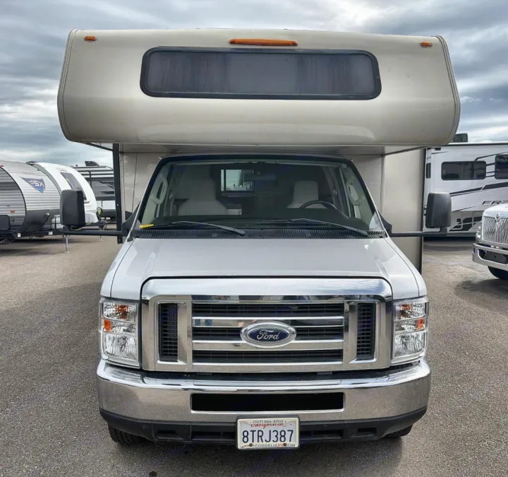 Front view of a white Ford Class C RV rental, parked outdoors under an overcast sky, with other RVs in the background.. Coachman Leprachaun 2020