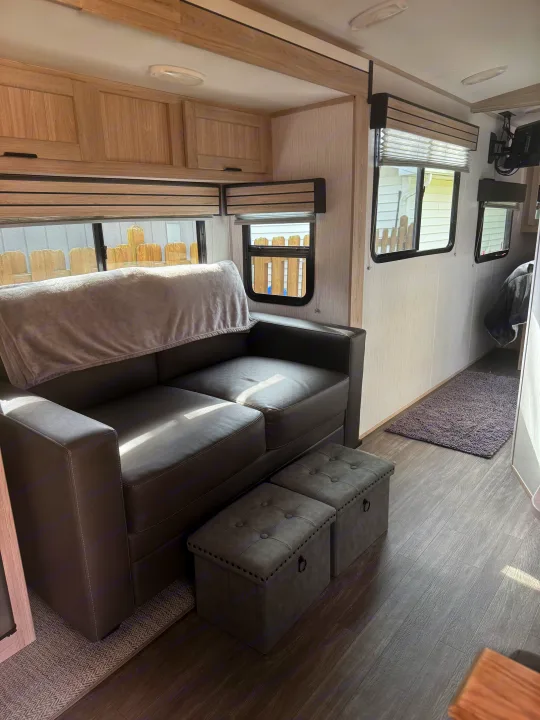 Modern RV rental interior with a cozy grey sofa, storage ottomans, wood cabinetry, and large windows.. Cruiser RV Shadow Cruiser 2022