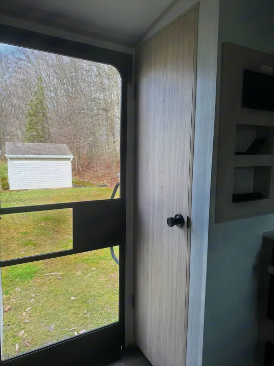 Inside an RV rental, an open screen door reveals a yard, shed, and bare trees. To the right: a wooden interior door and control panel.. Cruiser RV Shadow Cruiser 2022